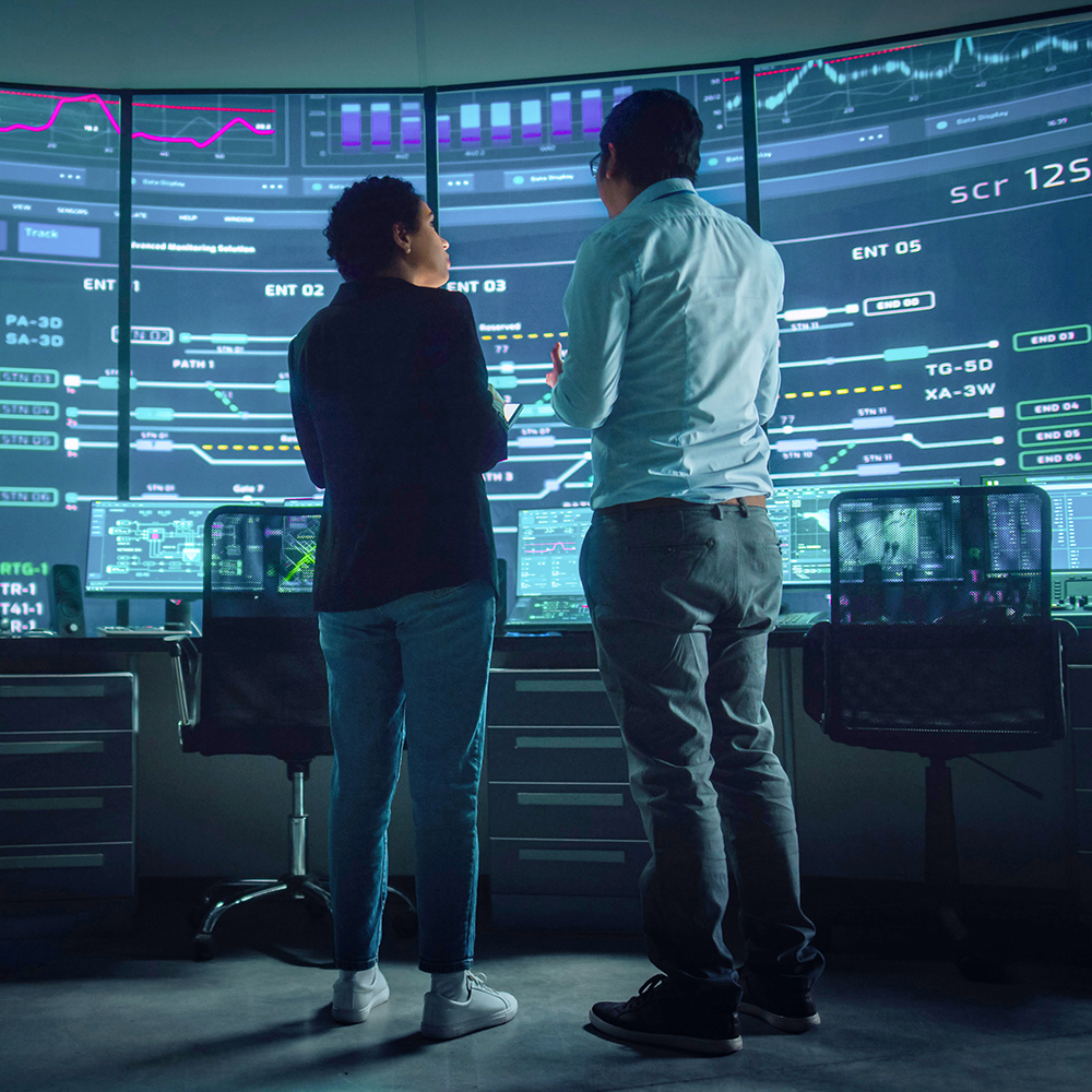 In the System Control Room Project Manager and IT Technical Engineer Have Discussion, they're surrounded by Multiple Monitors with Graphics